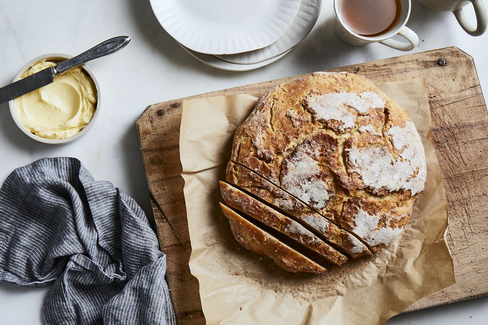 A sliced sourdough loaf on a wooden board with butter and tea